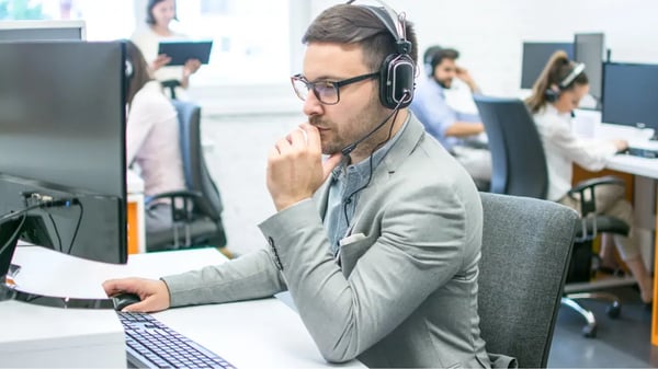 Profesional de atención al cliente con auriculares y micrófono trabajando frente a un ordenador en una oficina moderna, mientras otros agentes atienden llamadas al fondo. Profesional de atención al cliente con auriculares y micrófono trabajando frente a un ordenador en una oficina moderna, mientras otros agentes atienden llamadas al fondo.