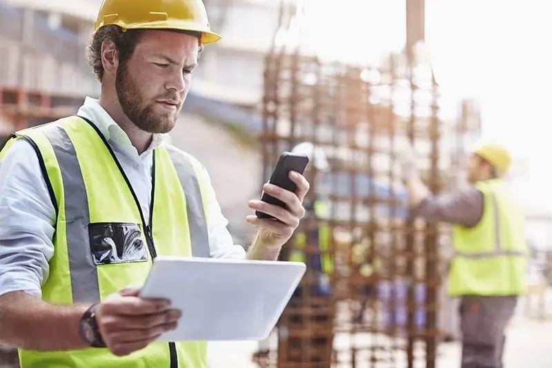 Man in a construction site checking his phone with a tablet on the other hand