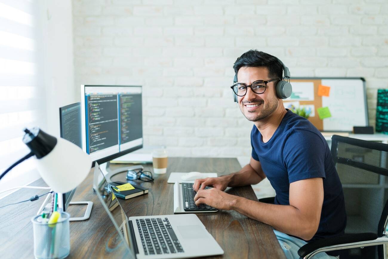 A smiling developer, coding in multiple screens on a desk