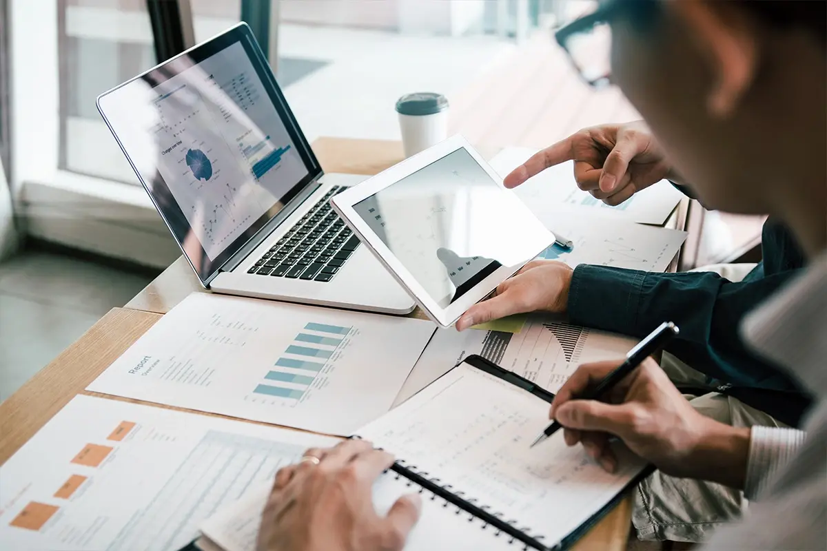 Person looking at a tablet while writing on a notebook on a desk with business related documents